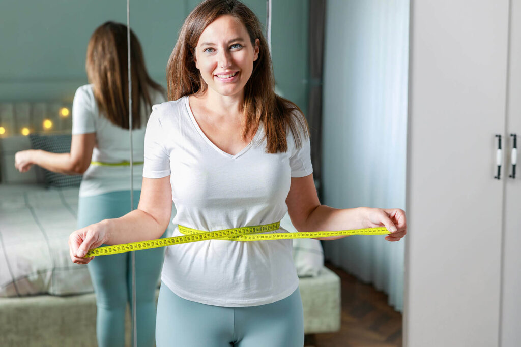 Smiling young woman after weight loss measuring waist in front of mirror