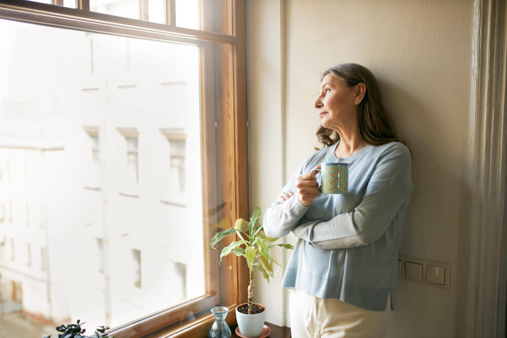 mature woman in casual clothes standing by large window,