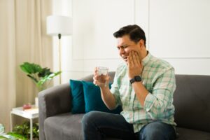 Male experiencing sharp tooth pain, gripping cold water glass while seated on living room