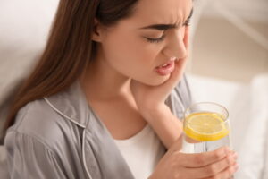 Young woman in pajamas with lemon water suffering from toothache at home, closeup