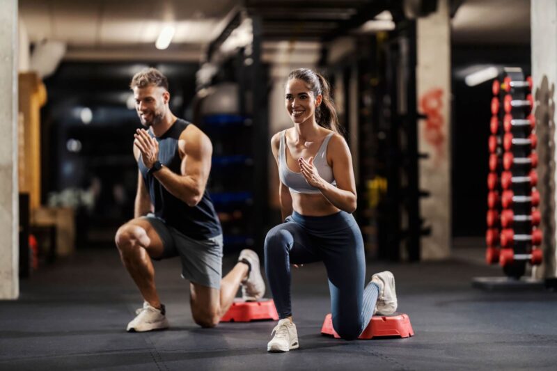 Happy sports people in shape are doing lunges in a gym