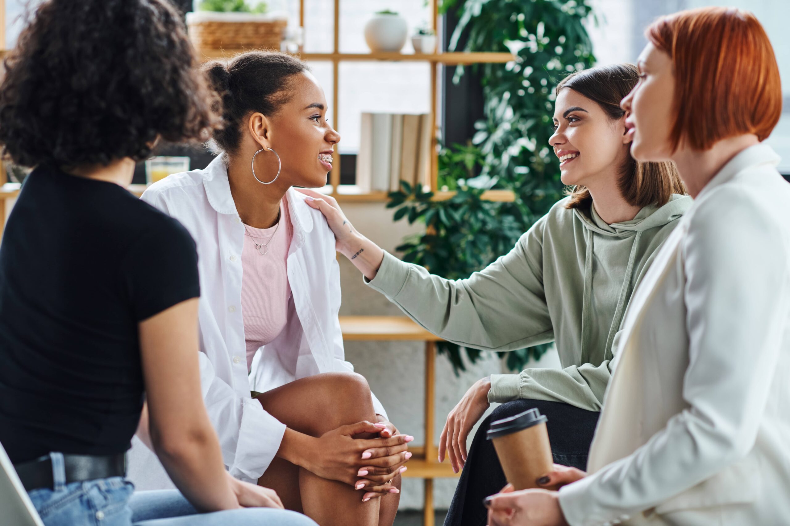 Young woman touching shoulder of pleased woman while sitting next to psychologist