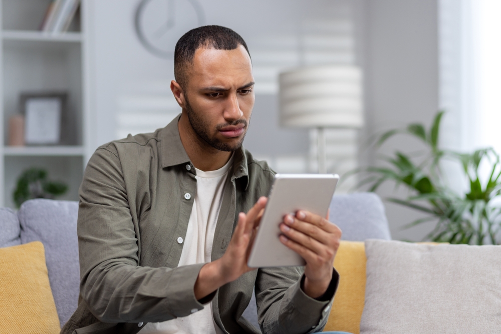 Worried young hispanic man sitting on sofa at home and using tablet. Saddenedly reads the news, checks the mail, received a message.