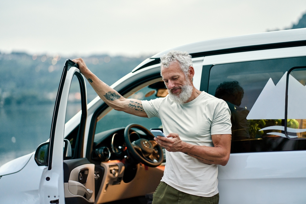 Mature tattooed man standing near rv camper van on vacation using mobile phone