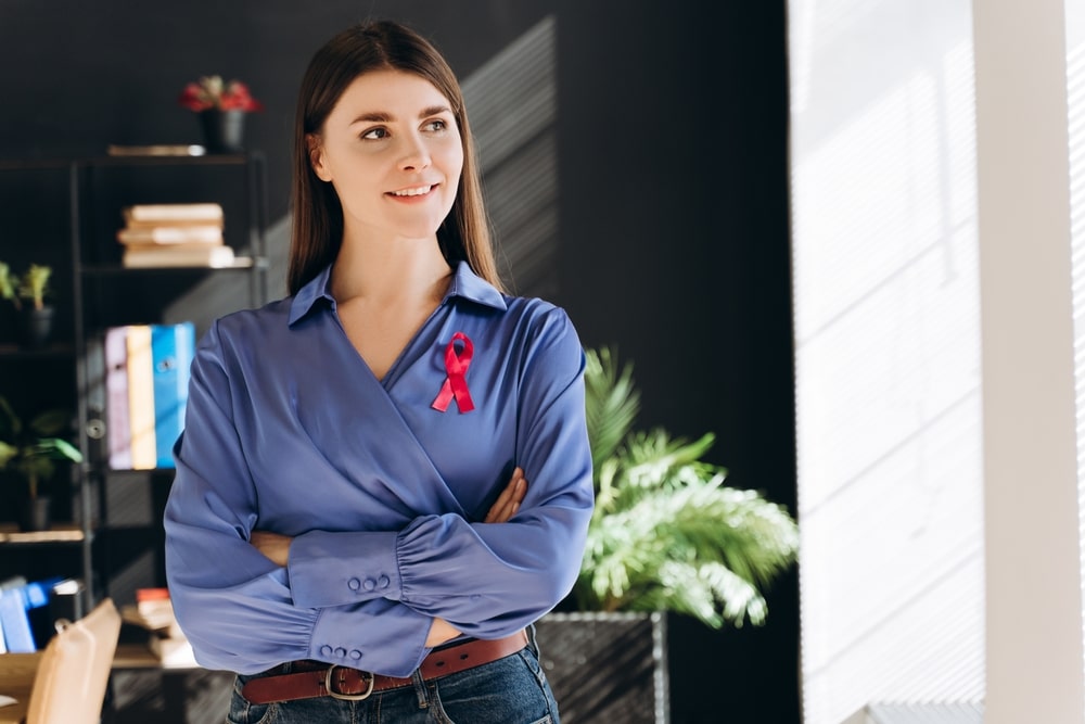 Young businesswoman wearing a red ribbon, smiling confidently with arms crossed, looking away in an office setting,
