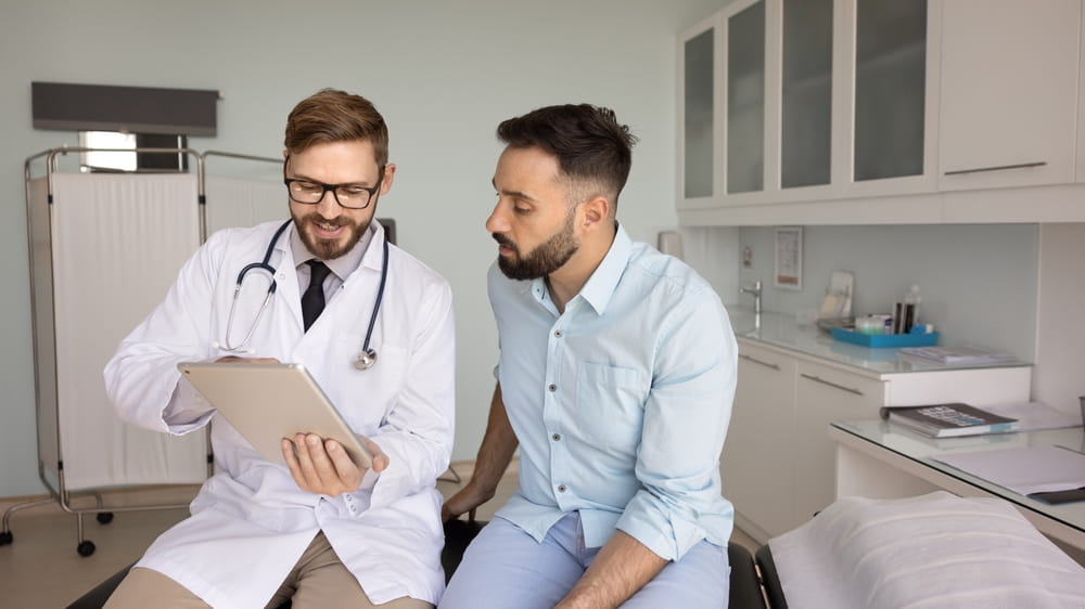 Positive young male doctor showing tablet to patient in exam room
