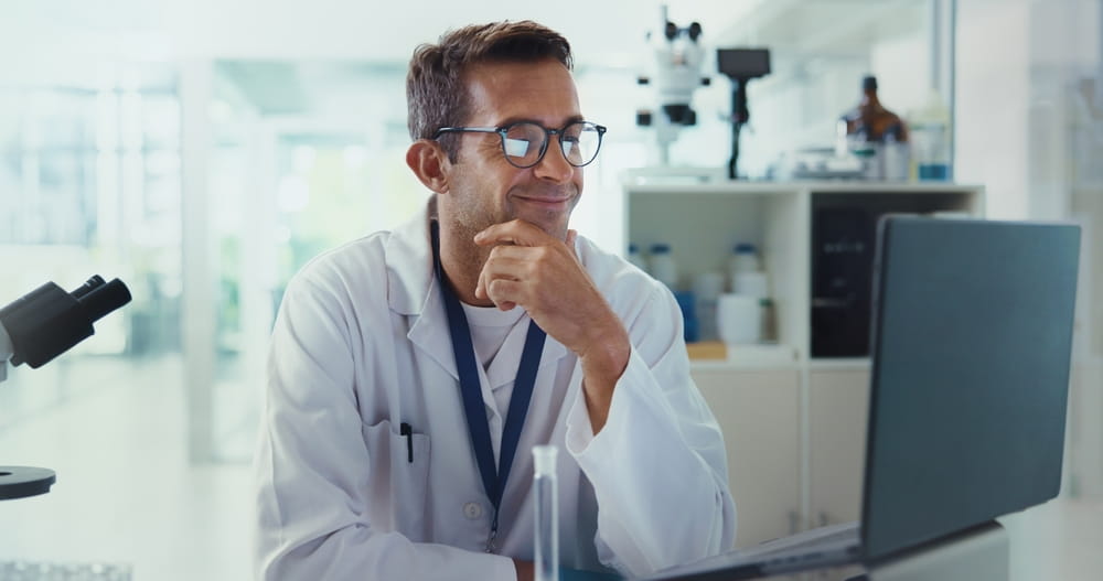Scientist, man and happy by laptop in lab for medical research with reading, review and vaccine development
