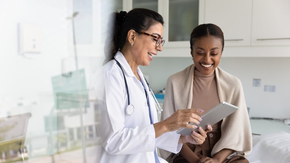 Happy senior practitioner woman and cheerful young African patient woman watching examination medical report on tablet computer