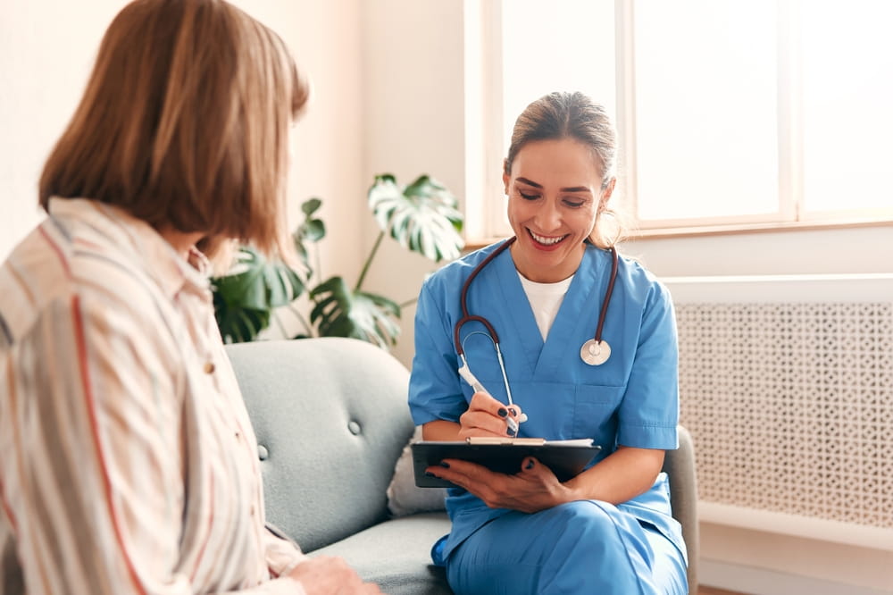 Happy young female nurse provide care medical service help support smiling mature woman at homecare medical visit,