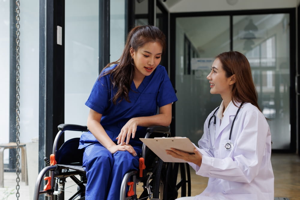 friendly female nurse staff caregiver of nursing home talking to asian young woman a wheelchair in hallway