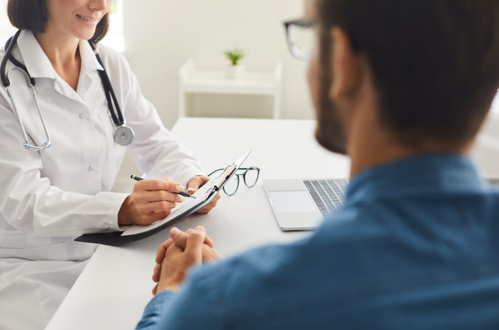 Woman who works as physician sitting at desk, talking to young man, writing down patient's prescription, giving advice and recommendations