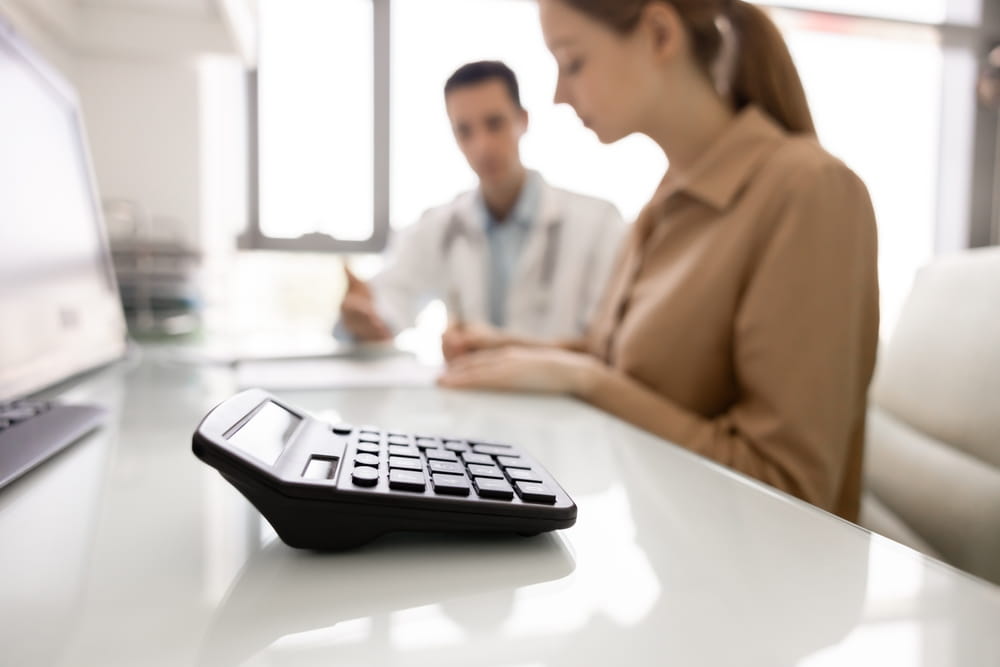 Close-up calculator on desk with blurred male doctor and female patient discussing medical paperwork in clinic.