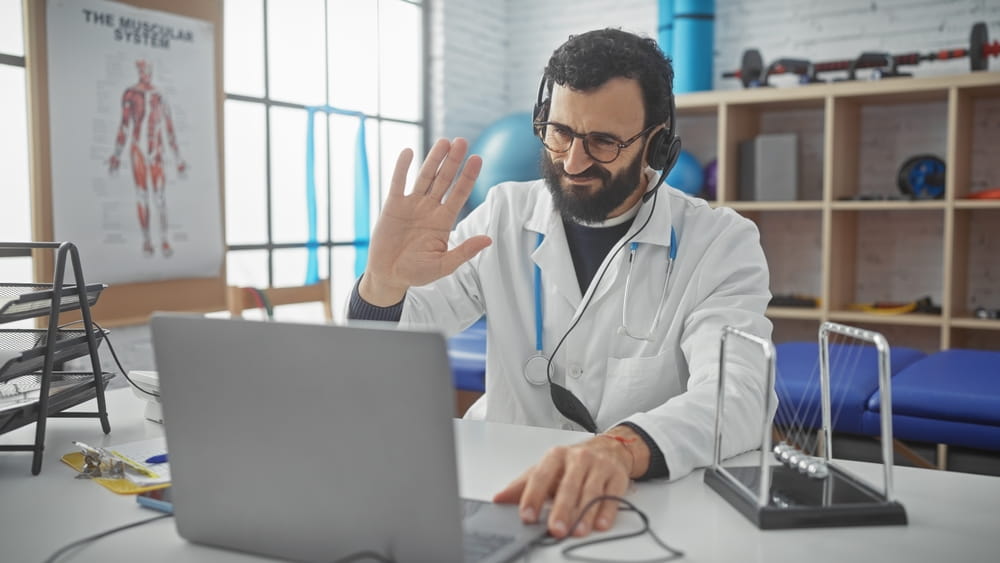 A bearded middle-aged male doctor in a white coat engaging in a telehealth consultation indoors at a clinic.