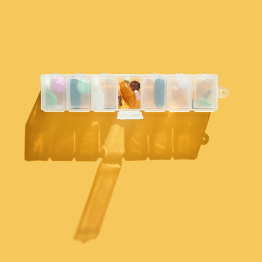 Weekly pill organizer with shadow on yellow background containing various colorful tablets.
