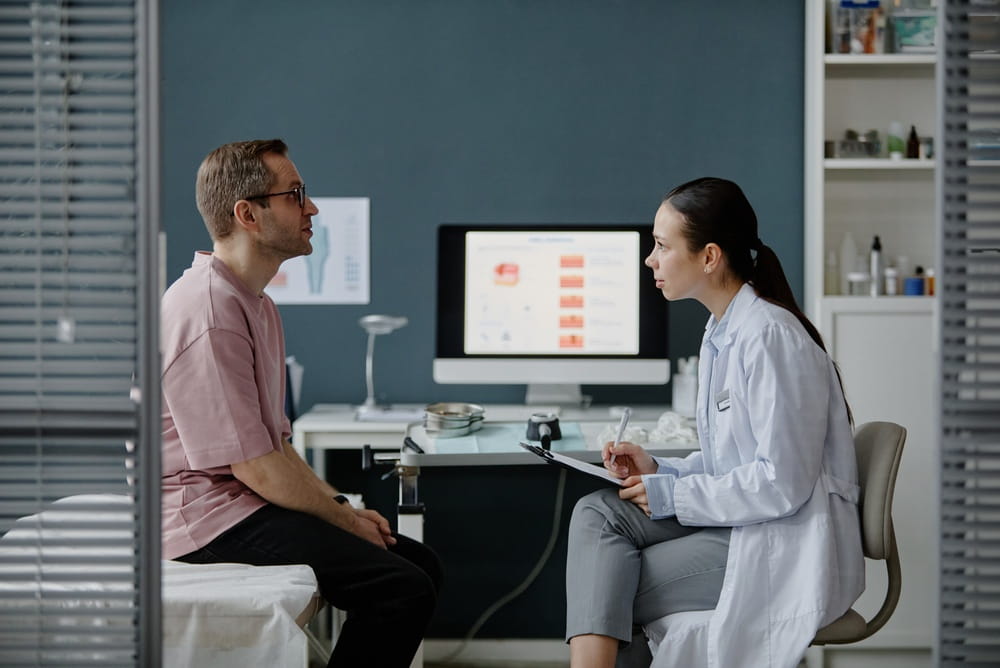 Caucasian middle aged man sitting on examination table consulting with Asian young adult woman dermatologist holding clipboard,