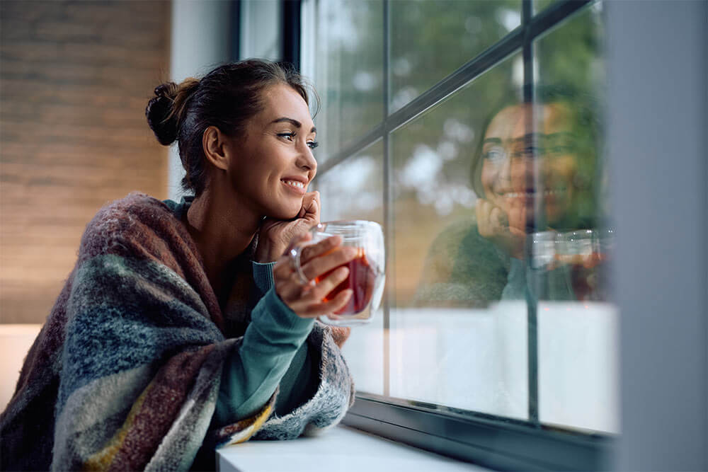 Young smiling woman drinking tea and looking through the window