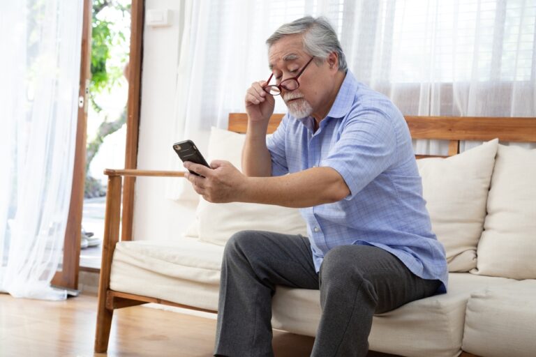 Senior Asian man moves his reading glasses to look at mobile phone while sitting on the living room couch at home