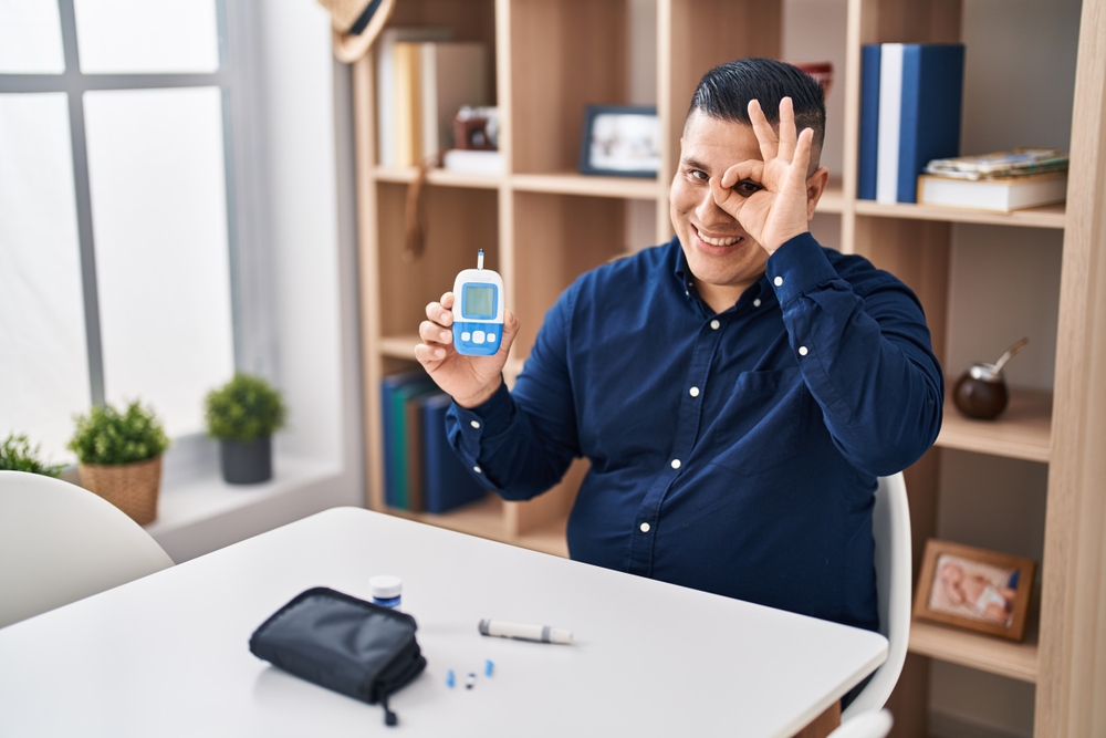 Hispanic young man holding glucometer device smiling happy doing ok sign with hand on eye looking through fingers