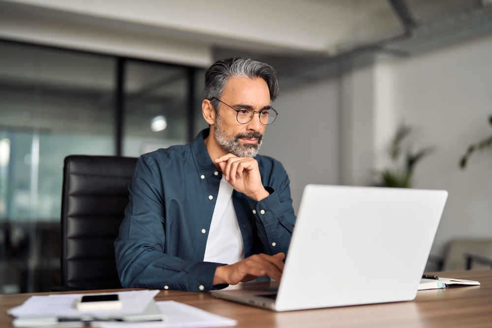 Busy middle aged business man executive wearing shirt and glasses sitting at desk using laptop