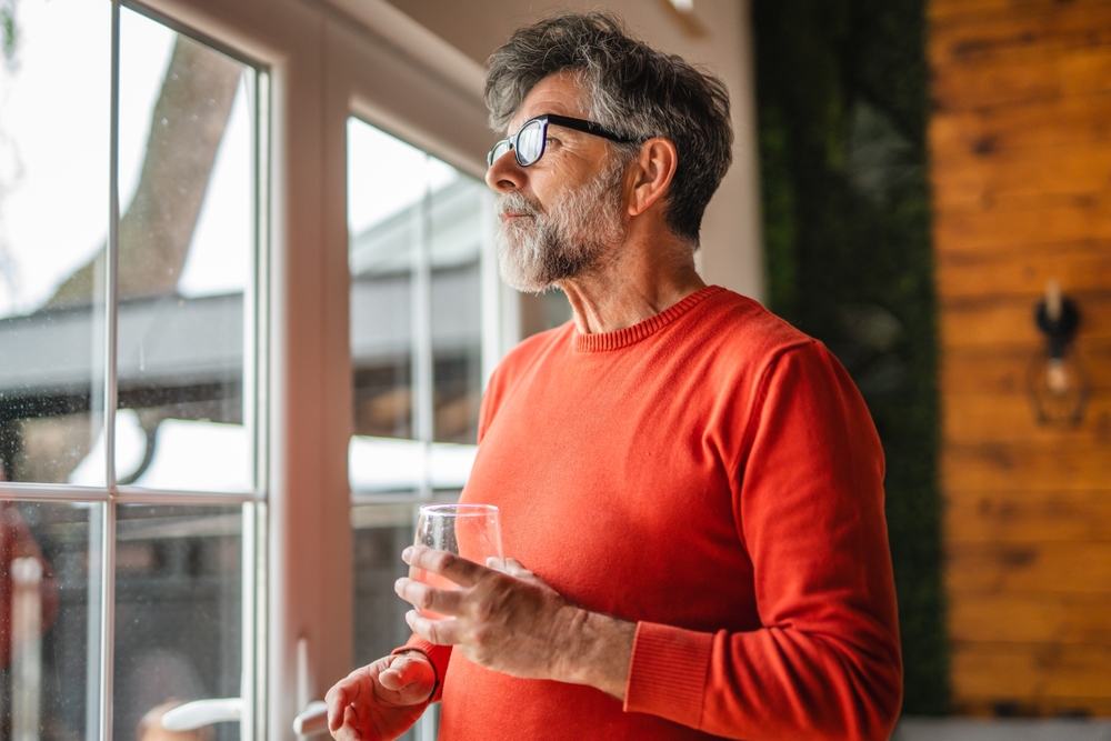 Portrait of aged man stand with glass of water and look through the window