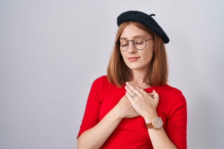 Young redhead woman standing wearing glasses and beret smiling with hands on chest with closed eyes and grateful gesture on face