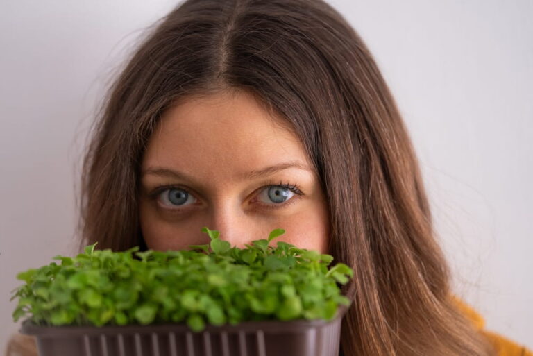 Close-up of woman with bright blue eyes holding pot of fresh microgreens in front of face
