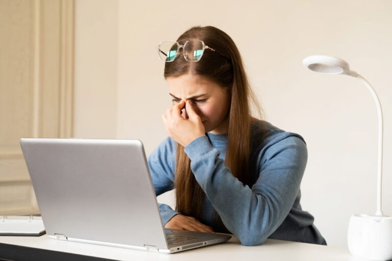 Tired young woman sitting at work with a laptop, rubbing the bridge of her nose due to eye strain fatigue