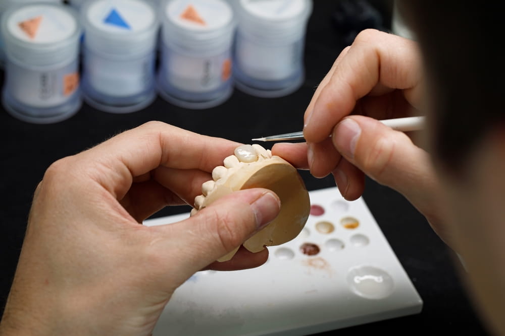 A dental technician applies ceramic to a crown using a brush.