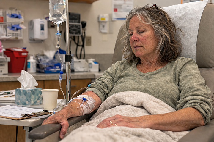 Woman with should-length gray hair seated in medical chair, covered in blanking, with IV hooked up to her right arm.