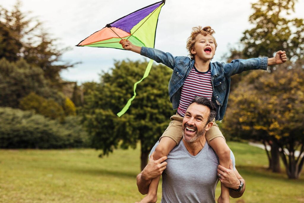 Smiling boy playing with colorful kite outside sitting on father's shoulders