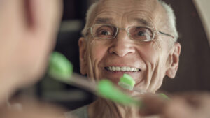 Gray haired senior man is cleaning his teeth and smiling looking in mirror in bathroom