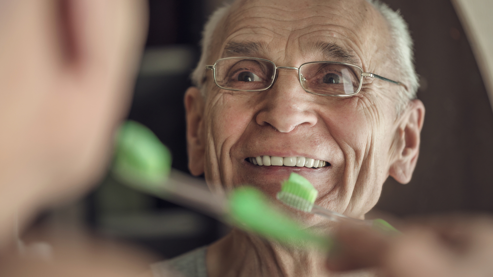 Gray haired senior man is cleaning his teeth and smiling looking in mirror in bathroom