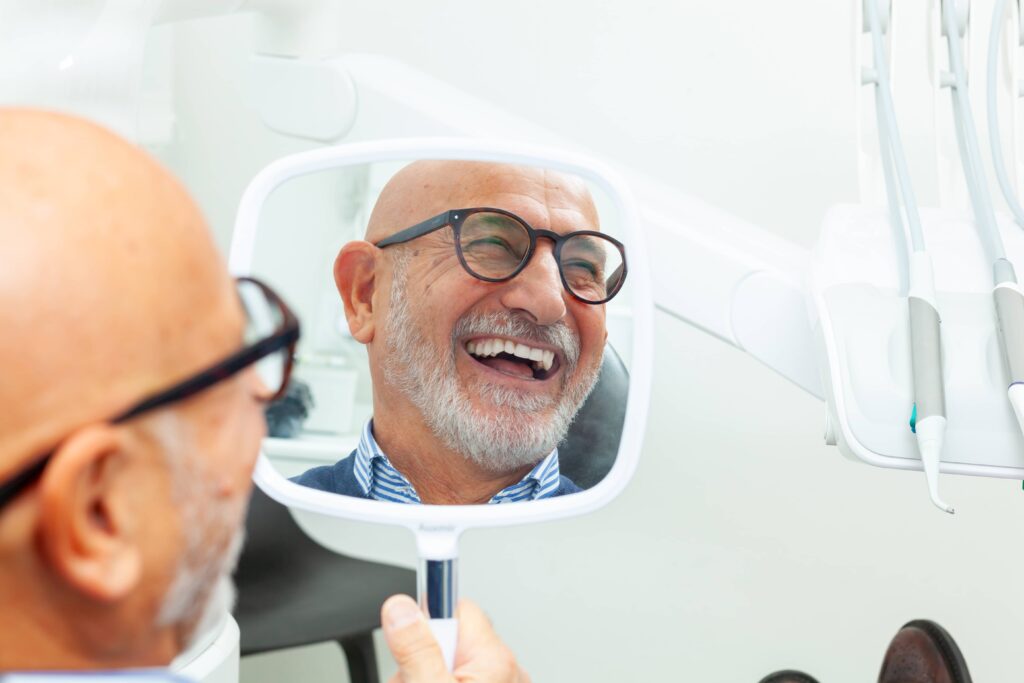 Happy senior patient admiring his perfect smile after a dental implant treatment, holding a mirror