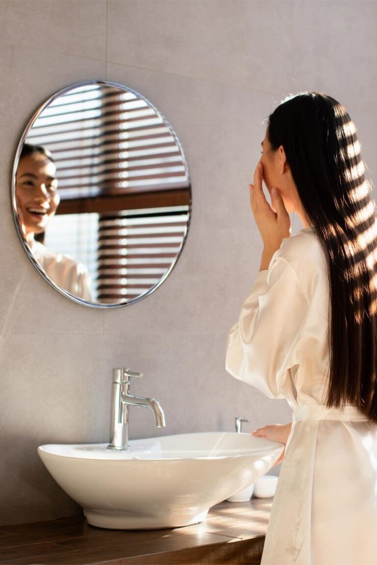 Beautiful long-haired young lady in white bathrobe looking at mirror