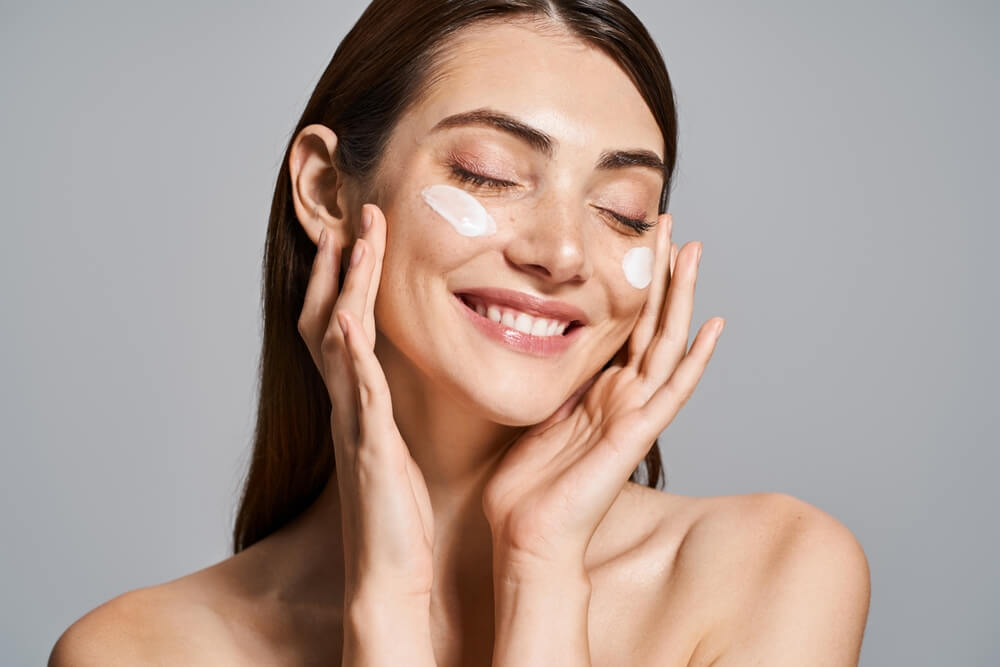 Woman smiles brightly while delicately touching her face with cream