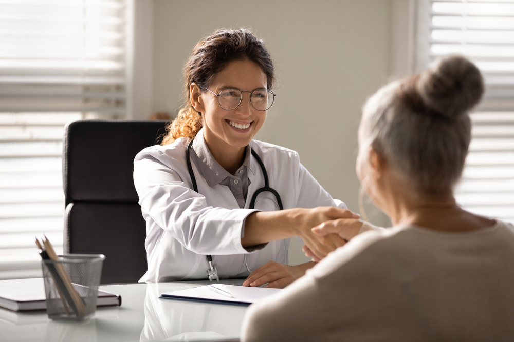 Close up smiling female doctor wearing glasses and uniform with stethoscope shaking mature woman hand at meeting, elderly patient
