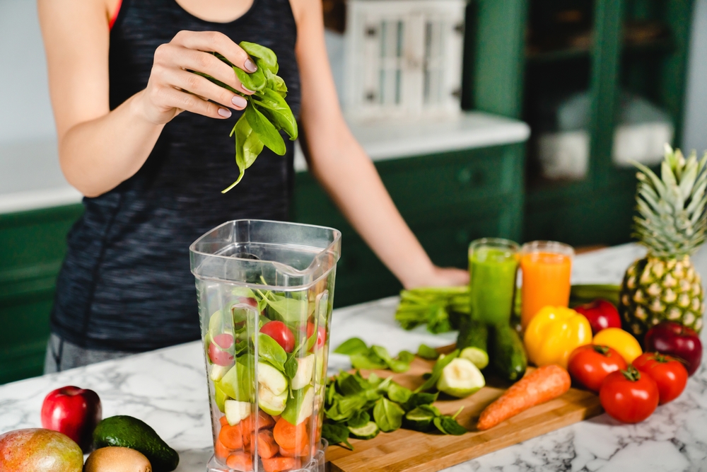 Young girl making green detox cocktail of fresh vegetables and fruits using blender in the kitchen