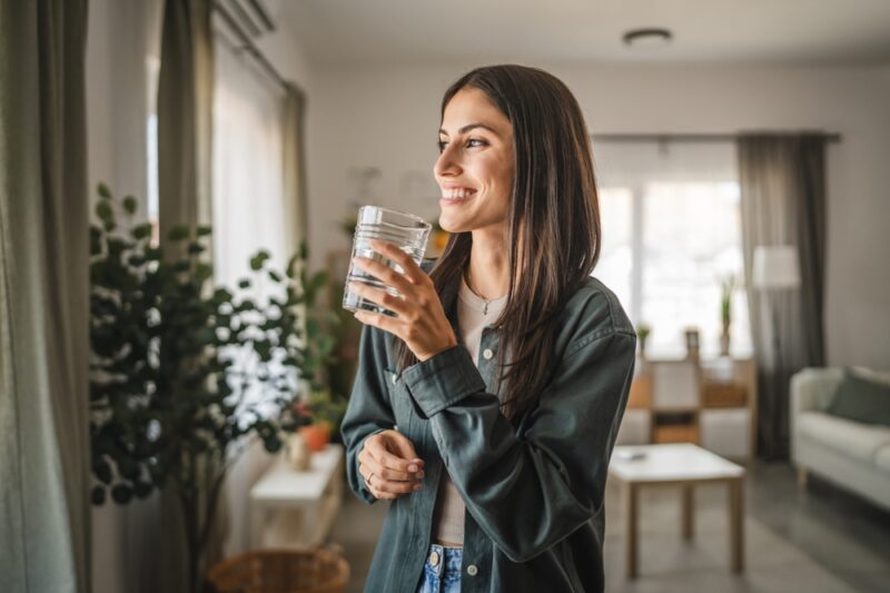 Portrait of adult women stand hold glass of water and look out the window