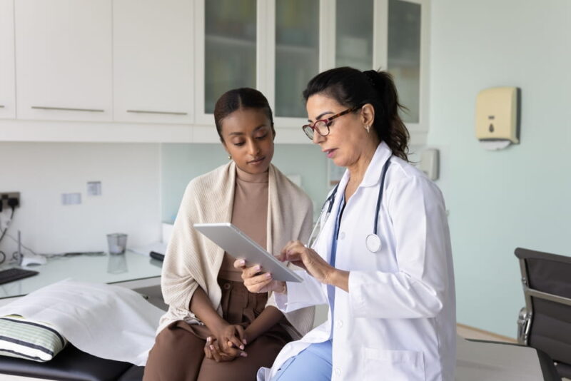 Elderly 60s doctor using digital tablet, showing medical scans or health lab test results to African woman patient at visit in clinic office