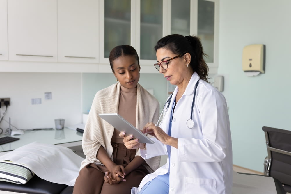 Elderly 60s doctor using digital tablet, showing medical scans or health lab test results to African woman patient at visit in clinic office