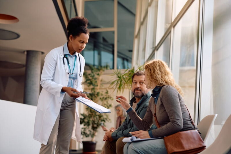 African American doctor talking to a couple while going through their medical documents in waiting room at the clinic.