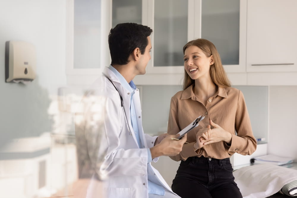 Happy young 20s female patient talking to male physician in examination room, smiling while doctor filling hospital form, writing medical records, discussing healthcare and treatment