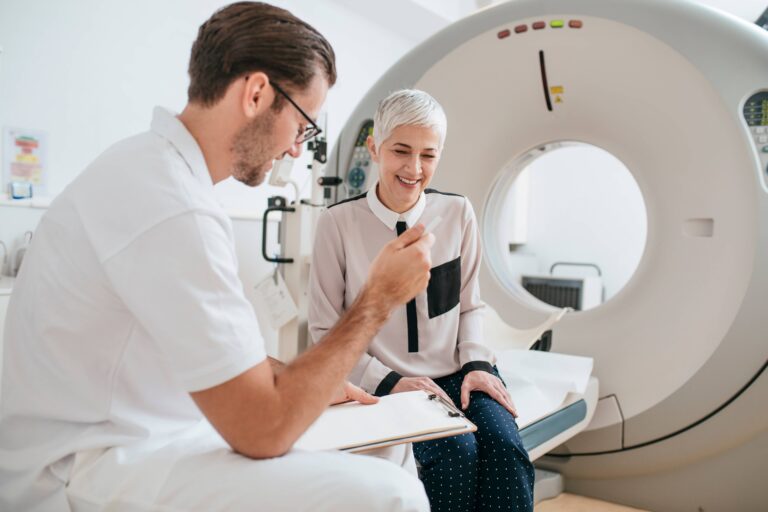Young medical technician talking to a patient and taking notes while sitting on the CT scanner bed.