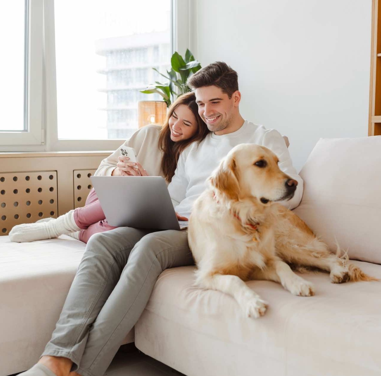 Portrait of happy young couple using laptop, man holding mobile phone, hugging dog, sitting together on comfortable sofa at home.