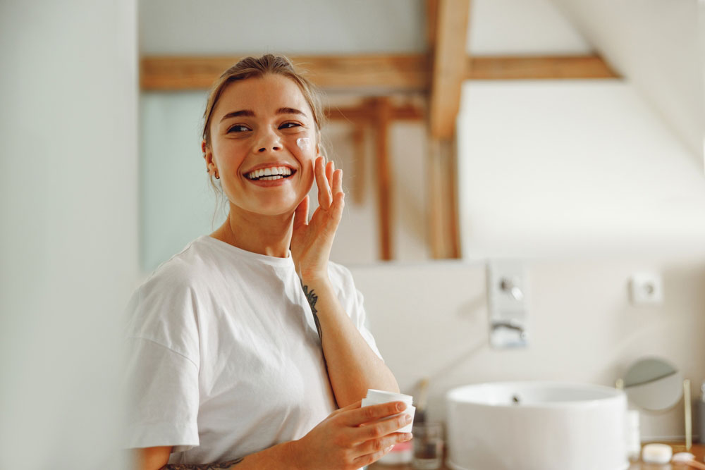 Beautiful young woman taking care of skin by applying moisturizer cream