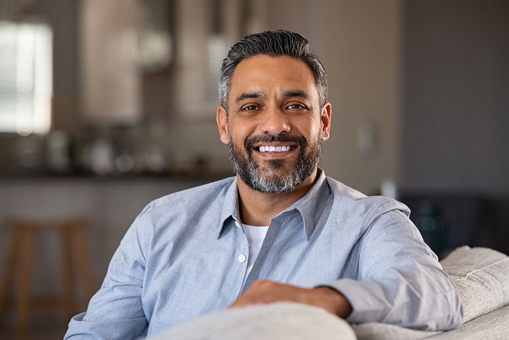 Portrait of happy mid adult man sitting on sofa at home. Handsome latin man in casual relaxing on couch and smiling