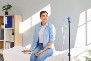 smiling woman sitting on the couch in medical clinic looking at the window while receiving IV drip infusion and vitamin injection therapy in her blood