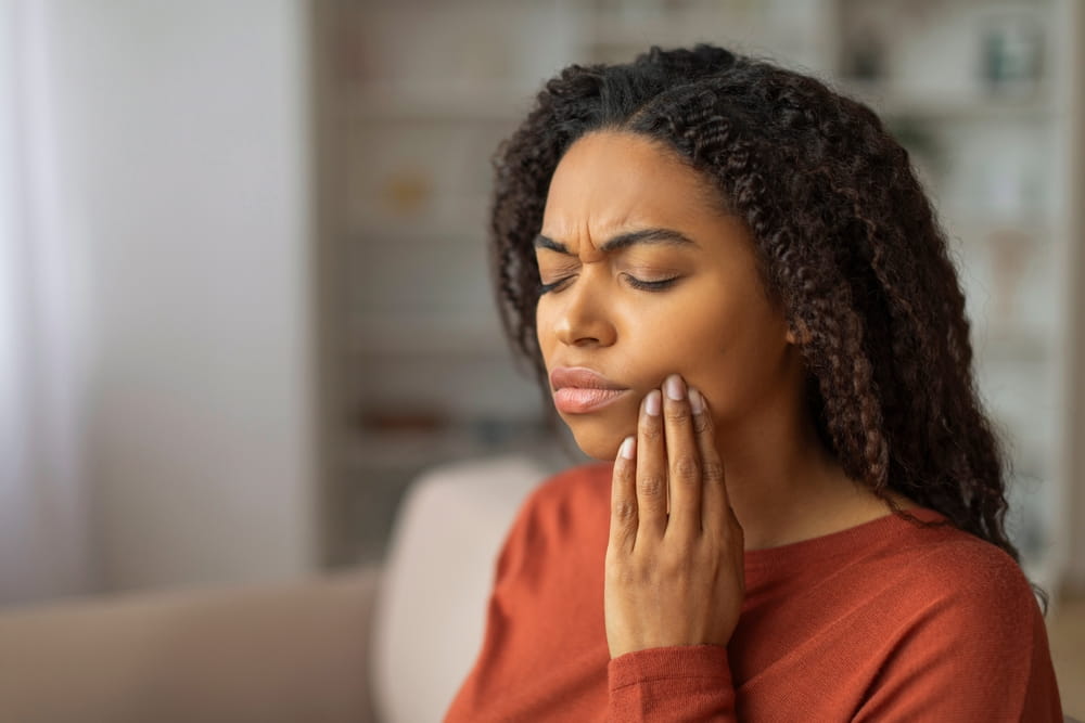 A young black woman pressing her cheek, showing discomfort from a possible toothache, african american female feeling unwell at home