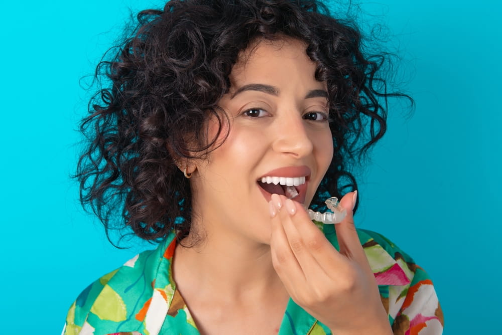 young arab woman wearing colorful shirt over blue background holding an invisible aligner ready to use it
