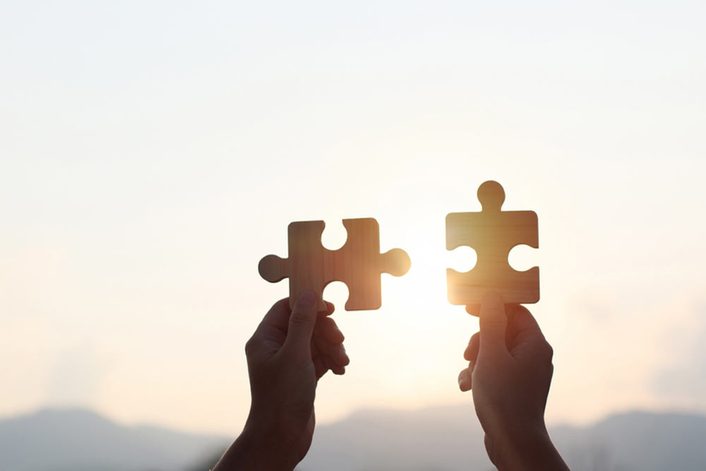 Hands are raised against a bright, white sky, each holding a piece of a wooden jigsaw puzzle. teamwork